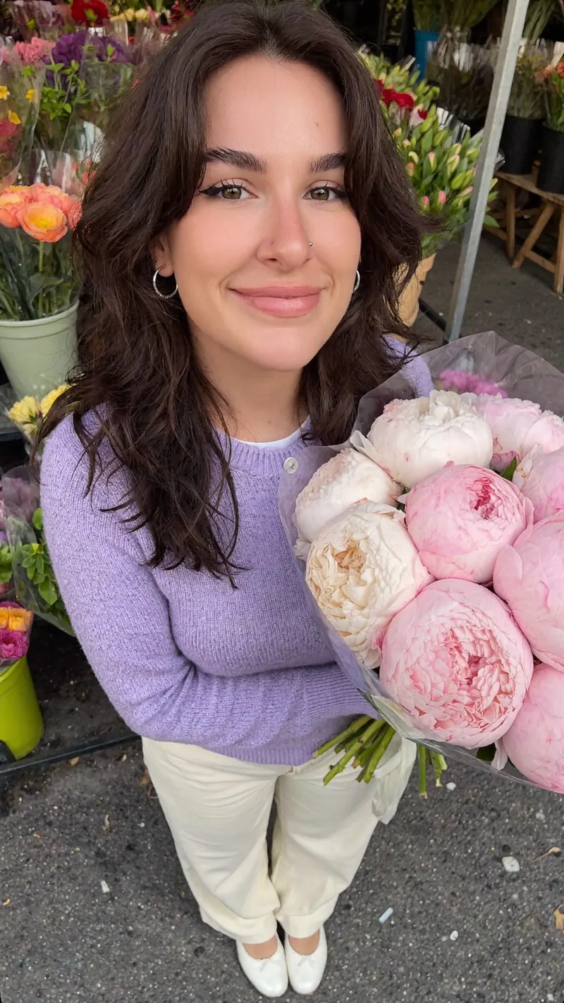 Olivia smiling at a flower market in a lilac sweater, holding a bouquet of pink peonies
